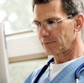 Arzt in blauer Krankenhauskleidung und Stethoskop am Laptop.
