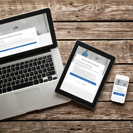 Laptop, Tablet und Handy vor Holzhintergrund Auf dem Bild sind ein eingeschalteter Laptop mit einem Tablet sowie einem Smartphone auf einer Holzoberfläche dargestellt.