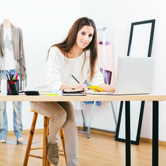 Frau am Schreibtisch Das Bild zeigt eine Frau, die auf einem Hocker an einem Schreibtisch sitzt und einen Stift in der Hand hält. Vor ihr steht ein Laptop.
