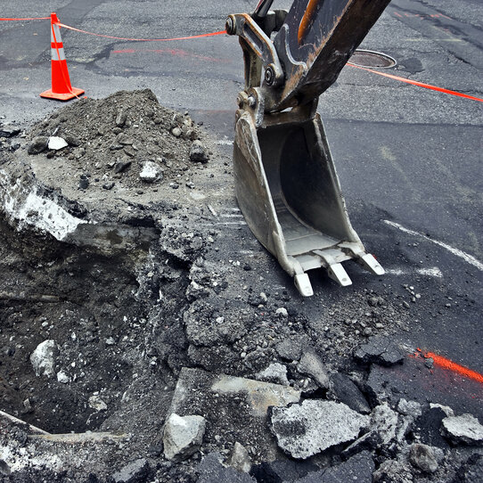 Baggerschaufel Baggerschaufel neben einem ausgeschaufelten Loch im Asphalt, daneben Pylonen.
