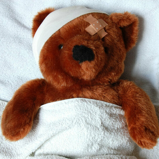Verletzter brauner Tedddybär mit einem Verband und einem Pflaster am Kopf, zugedeckt in weißen Laken liegend.
