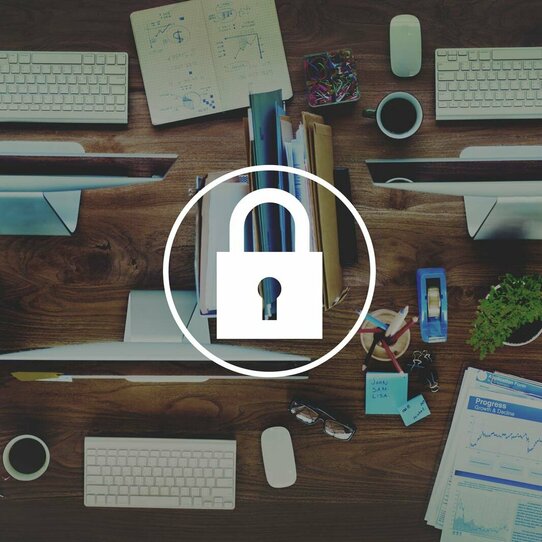 IT-Sicherheit Versinnbildlichung von IT-Sicherheit anhand eines Vorhängeschloss-Ikons vor einer Draufsicht auf Computer mit Tastaturen sowie Schreibtisch-Utensilien.