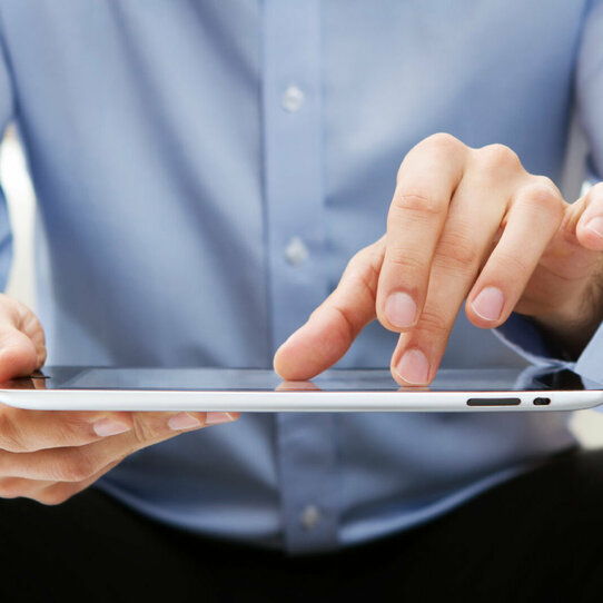 Person am Tablet Das Bild zeigt einen Ausschnitt einer Person mit blauem Hemd und schwarzer Hose, die ein Tablet nutzt.