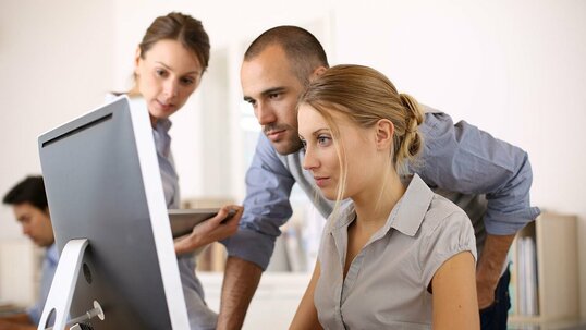 Drei Personen, zwei Frauen und ein Mann, die sich gemeinsam etwas an einem Computer-Monitor ansehen. 