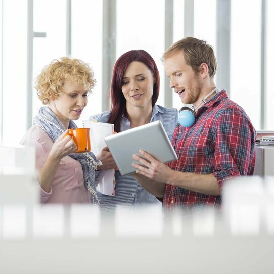 Bürosituation mit einem Mann und zwei Frauen Das Bild zeigt einen Mann und zwei Frauen im Gespräch über etwas, was auf dem Tablet dargestellt wird.