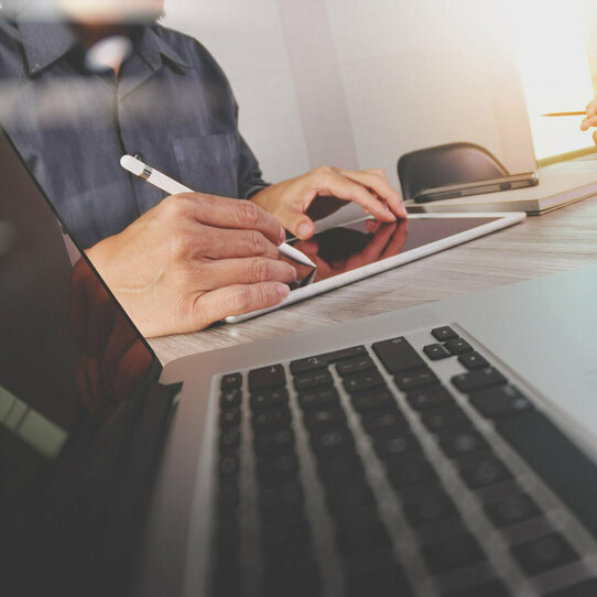Arbeitssituation mit Laptop, Tablet und Handy Zwei Arbeitskollegen, nur ausschnittweise zu sehen, arbeiten mit einem Tablet, Handy und Laptop an einem Tisch.