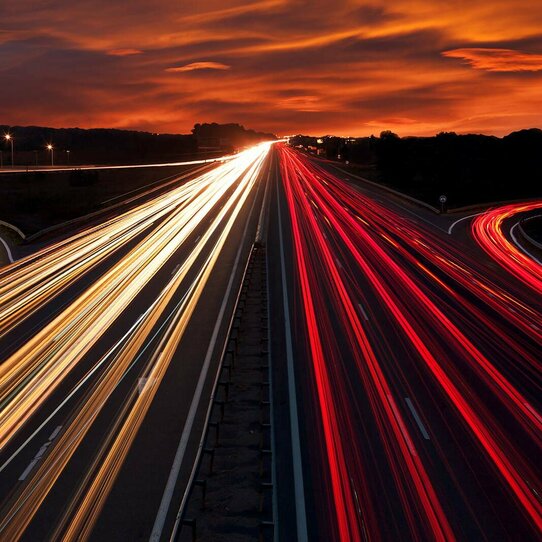 Autolichter auf Autobahn bei Nacht Langzeitbelichtung: Rote und weiße Streifen von Autolichtern auf einer Autobahn vor orangefarbenem Himmel.