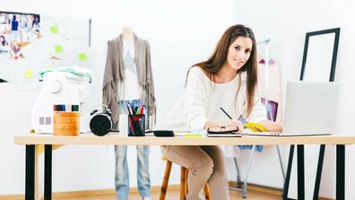 Das Bild zeigt eine Frau, die auf einem Hocker an einem Schreibtisch sitzt und einen Stift in der Hand hält. Vor ihr steht ein Laptop.