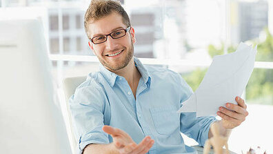Das Bild zeigt einen lächelnden Mann mit Brille in einem hellblauen Hemd. Er sitzt vor einem Monitor  eine Hand streckt er der Kamera entgegen, in der anderen hält er mehrere Blätter Papier. 