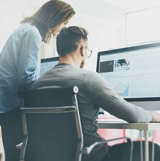 Mitarbeiter am Schreibtisch Das Bild zeigt eine alltägliche Bürosituation. Ein Mitarbeiter sitzt an seinem Schreibtisch am Computer, eine weitere Person steht daneben. Beide betrachten die Darstellung auf dem Monitor. Im Hintergrund sind verschwommen weitere Personen zu erkennen.