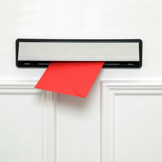 Das Bild zeigt einen roten Briefumschlag, der aus einem Briefschlitz in einer weißen Tür herausragt. 