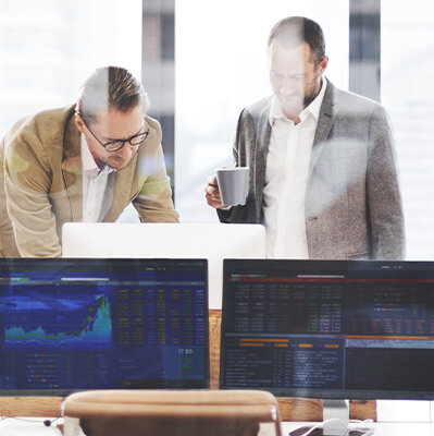 Zwei Männer vor Monitor Arbeitssituation im Büro, bei der zwei Männer vor einem Computermonitor stehen. Im Vordergrund sind weitere, eingeschaltete Monitore zu erkennen.