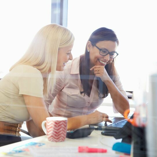 Zwei Frauen sitzen gemeinsam am Schreibtisch und arbeiten an einem Laptop. 
