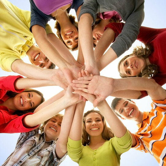 Personengruppe mit in der Mitte zusammengelegten Händen Gruppe von bunt bekleideten Personen, die ihre Hände in der Mitte zusammenlegen und in die Kamera lächeln. Von unten aufgenommen.