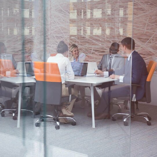 Geschäftsleute an Schreibtisch Mehrere Geschäftsleute an einem Schreibtisch in einem modernen Büro. Das Foto wurde durch eine Glastür aufgenommen, die im Vordergrund sichtbar ist.