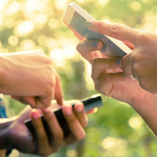 Hände mit Smartphones Nahaufnahme den Händen zweier Personen, die ihre Smartphones benutzen. Im Hintergrund sind verschwommen Bäume und Sonnenschein zu erkennen.