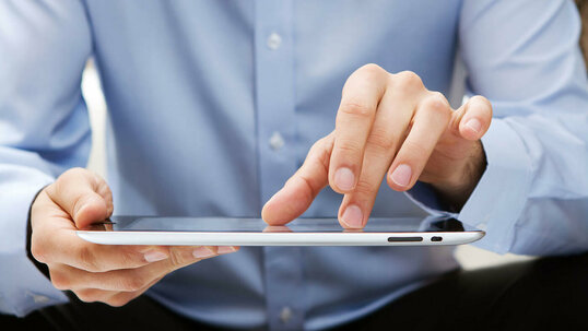 Das Bild zeigt einen Ausschnitt einer Person mit blauem Hemd und schwarzer Hose, die ein Tablet nutzt.