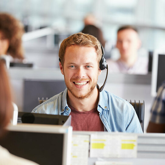 Mitarbeiter mit Headset Mann mit Headset lächelt in die Kamera. Im Hintergrund sind weitere Kollegen zu erkennen, diese sind aber verschwommen.