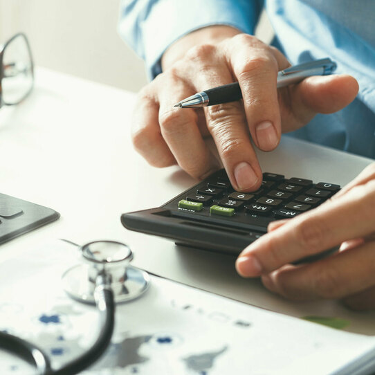 Das Bild zeigt ausschnittsweise eine Person am Taschenrechner sowie ein Stethoskop, einen Laptop und eine Brille. Bildliche Darstellung der Kostenberechnung im Gesundheitswesen.