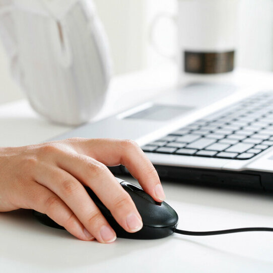 Das Bild zeigt den Ausschnitt einer weiß gekleideten Person am Schreibtisch vor einem Laptop mit Fokus auf die Hand auf der Computermaus.