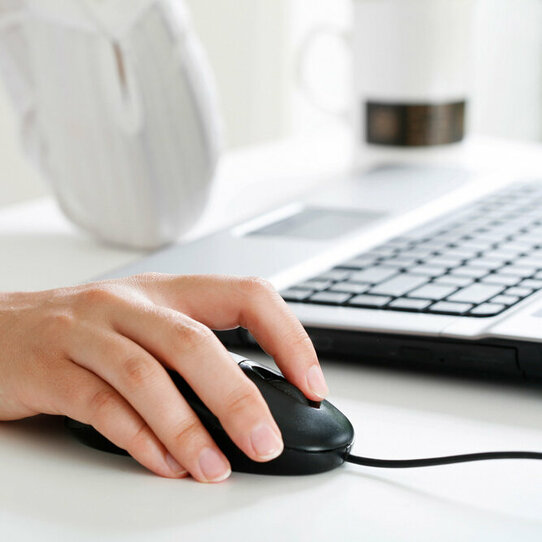 Hand auf Computermaus Das Bild zeigt den Ausschnitt einer weiß gekleideten Person am Schreibtisch vor einem Laptop mit Fokus auf die Hand auf der Computermaus.