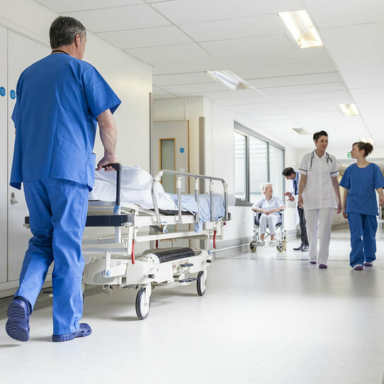 Das Bild zeigt einen Pfleger, der ein Krankenbett durch einen Krankenhausflur schiebt. In dem Flur befindet sich noch weiteres medizinisches Personal, das ihm entgegen kommt.