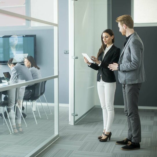 Zwei Personen mit Tablet im modernen Büro Mann und Frau stehen vor einem modernen Büro und betrachten etwas auf dem Tablet. Im Hintergrund sitzen zwei Personen an einem Besprechungstisch.