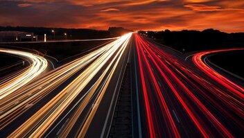 Langzeitbelichtung: Rote und weiße Streifen von Autolichtern auf einer Autobahn vor orangefarbenem Himmel.