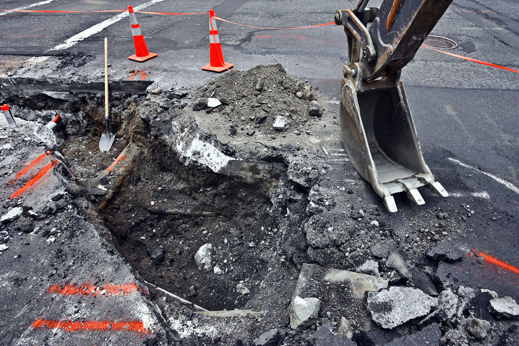 Baggerschaufel Baggerschaufel neben einem ausgeschaufelten Loch im Asphalt, daneben Pylonen.